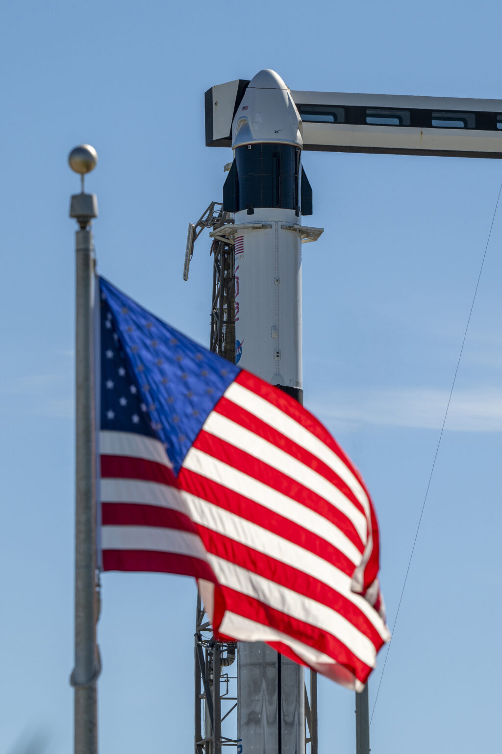 A close-up view of a SpaceX Crew Dragon spacecraft mounted atop a Falcon 9 rocket on the launch pad. The capsule and trunk are connected to ground support hardware, with NASA and mission insignia visible on the rocket’s exterior against a bright blue sky. in the foreground in the American flag.