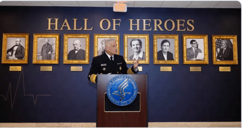 A uniformed official speaks at a podium in front of a “Hall of Heroes” wall, with framed portraits of notable individuals displayed behind him.