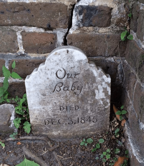A weathered, white marble tombstone sits against a cracked brick wall. The inscription reads: “Our Baby” followed by “DIED DEC. 3, 1845.” Small green weeds and leaves grow in the dirt around the base of the headstone.