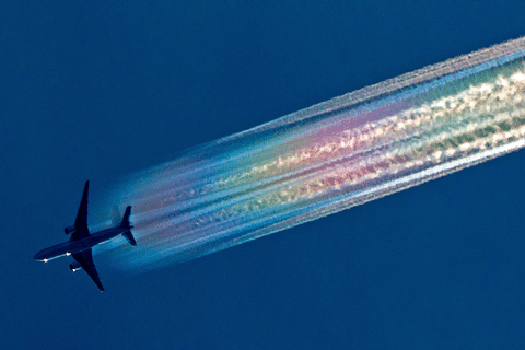 A commercial airplane in flight, leaving a bright, iridescent rainbow-colored contrail against a deep blue sky.