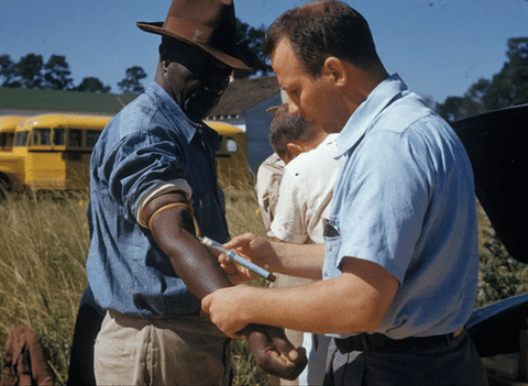 A man in a light blue shirt draws blood from the arm of a man wearing a hat, with a yellow school bus in the background.