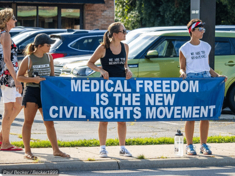 Three women holding a blue banner that reads, “MEDICAL FREEDOM IS THE NEW CIVIL RIGHTS MOVEMENT.”.