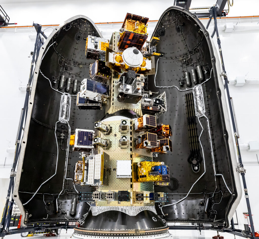 Top‑down view of a SpaceX payload fairing interior showing mounted satellites and hardware arranged along the center structure, surrounded by the curved black fairing walls.